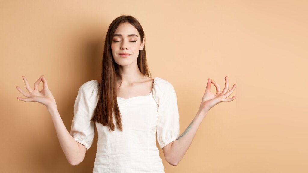 Junge Frau mit einer weißen Bluse hat die Augenschlossen und die Hände in Meditationshaltung und arbeitet an ihrer mentalen Gesundheit.