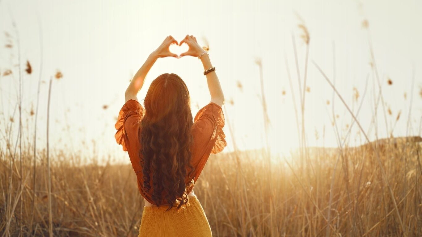 Stilvolle Frau vor einer Weide und dem Sonnenuntergang macht mit den Händen ein Herz über dem Kopf und möchte Selbstliebe lernen mit Anleitungen und Übungen.